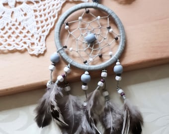 May include: A grey dreamcatcher with white and purple beads and brown feathers. The dreamcatcher is hanging from a white crocheted doily.