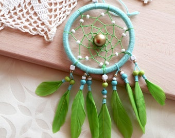 May include: A handmade dreamcatcher with a turquoise hoop, green and white web, and a gold bead center. The dreamcatcher features green feathers and decorative beads in shades of blue, green, and white. The craft is displayed on a wooden surface.