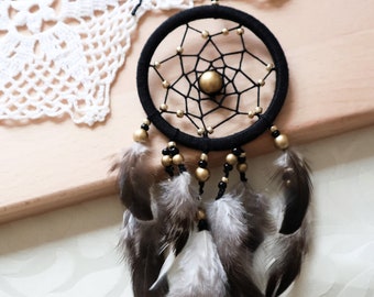 May include: A black dreamcatcher with a woven web design, gold accents, and hanging feathers. The dreamcatcher is suspended against a light background, showcasing its intricate details and natural aesthetic.
