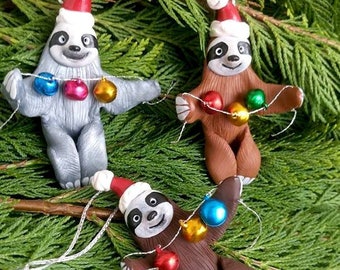 Puede incluir: Tres adornos navideños de perezosos. Cada perezoso lleva un gorro de Papá Noel y sostiene una cadena de luces navideñas de colores. Los perezosos son grises y marrones, con detalles blancos y ojos negros. Sobre un fondo de agujas de pino verdes.