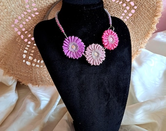 Peut inclure: Un collier floral avec trois pendentifs en forme de fleurs en tissu, dans des tons de rose et de violet. Le collier est présenté sur un buste de mannequin noir, avec un chapeau de paille en arrière-plan. Les fleurs ont un design détaillé et superposé.