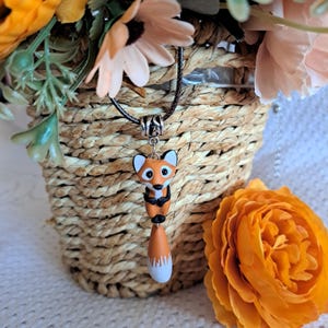 May include: A handcrafted fox pendant necklace. The pendant features a detailed fox with orange, white, and black accents. The fox is suspended from a black cord necklace. The background includes a woven basket and orange flowers.