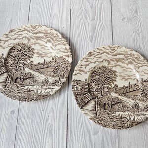 Può includere: Due piatti in ceramica marrone e bianco con un design di scena di campagna. I piatti presentano un ponte marrone, alberi ed edifici.