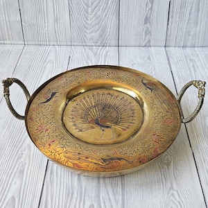 Puede incluir: Bandeja ovalada de latón con diseños grabados de pavo real y flores. Cuenta con dos asas ornamentadas. La bandeja es de color dorado cálido y se encuentra sobre una superficie de madera clara.