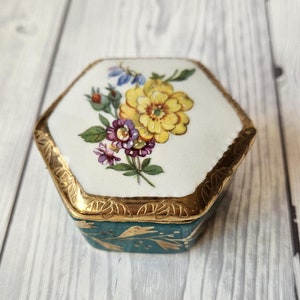 Könnte beinhalten: Eine kleine sechseckige Schmuckschatulle mit einem teelgrünen Sockel und einem weißen Deckel. Der Deckel ist mit einem Blumendesign verziert, das gelbe, violette und blaue Blumen zeigt. Die Schachtel hat einen goldenen Rand um die Kanten.