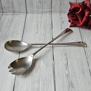 May include: A set of two silver salad serving utensils, a spoon and a fork, on a white wood background.
