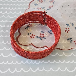 May include: A red beaded bracelet. The bracelet is made of small, round beads strung together to form a wide band. The beads are a vibrant shade of red. The bracelet is displayed on a light gray surface with a wave pattern.