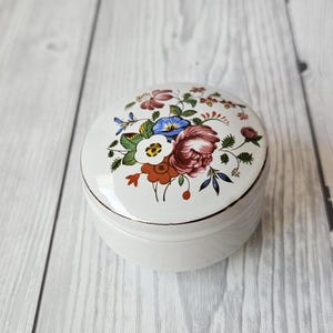 May include: A white ceramic trinket box with a floral design. The lid features a bouquet of pink, red, blue, and yellow flowers with green leaves.
