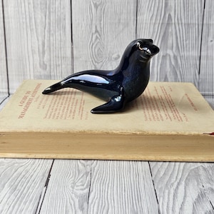 Könnte beinhalten: Eine dunkelblaue Glasrobbenfigur mit glänzendem Finish. Die Robbe liegt auf einem Vintage-Buch mit dem Titel "A Guide to Management Accounting". Die Figur ist ein dekorativer Artikel.
