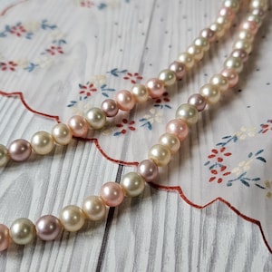 Könnte beinhalten: Eine zarte Perlenkette mit abwechselnd rosa- und cremefarbenen Perlen. Die Kette liegt auf einem weißen Stoff mit floralem Muster und rotem Wellenrand vor hellem Holzmaserungshintergrund.