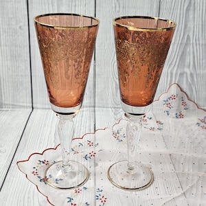 Könnte beinhalten: Zwei Vintage-Weingläser mit pfirsichfarbener Schale und Goldrand. Die Gläser haben einen klaren Stiel und Fuß. Sie stehen auf einer weißen Serviette mit rotem, gewelltem Rand und Blumenmuster. Der Hintergrund ist helles Holz.
