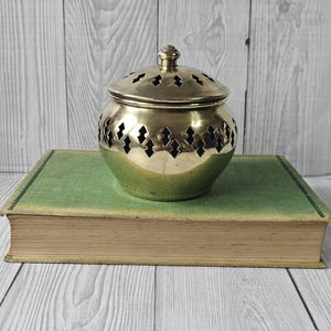 May include: A gold-coloured metal incense burner with a lid, featuring diamond-shaped cutouts. It sits atop a vintage green book with a worn cover, against a light grey wooden background.