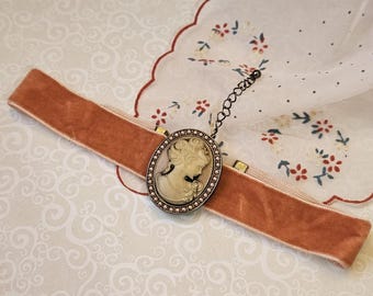Op de afbeelding: Een fluwelen choker in een gedempte oranje tint, met een camee hanger. De ovale hanger is omlijst met kleine, heldere stenen en een gedetailleerd profiel van een vrouw. De choker ligt op een patroon oppervlak.