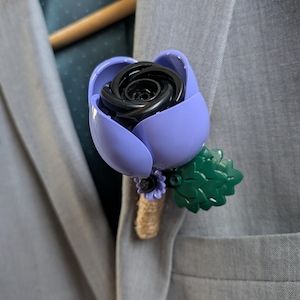Könnte beinhalten: Ein Ansteckblume bestehend aus einer schwarzen Rosenknospe in lavendelfarbenen Blütenblättern, akzentuiert mit einem kleinen grünen Blatt und einer winzigen violetten Blüte. Der Stiel ist mit beigefarbenem Garn umwickelt und am Revers eines hellgrauen Sakkos befestigt.
