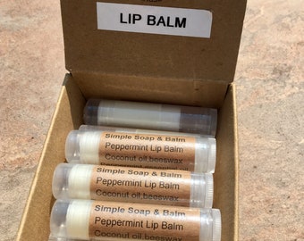 May include: A cardboard box containing several lip balm tubes. Each tube has a label that reads "Simple Soap & Balm Peppermint Lip Balm Coconut oil, beeswax". The box also has a label that says "LIP BALM". The background is a wooden surface.