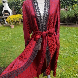 May include: A long, red robe with a floral pattern and black lace trim. The robe is tied at the waist with a matching belt. The robe is displayed on a mannequin in a garden setting.