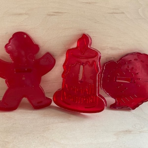 May include: Three red plastic cookie cutters: a gingerbread person, a birthday candle, and a turkey. The candle cutter has the words "Happy Birthday" on it.