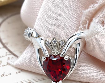 Könnte beinhalten: Ein Claddagh-Ring aus Silber mit einem herzförmigen, tiefroten Edelstein in der Mitte, der von zwei modellierten Händen gehalten wird. Über dem Herz befindet sich eine kleine Krone, die mit drei winzigen, klaren Steinen besetzt ist. Der Ring ist poliert.