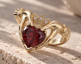 Könnte beinhalten: Ein goldener Claddagh-Ring mit einem herzförmigen roten Edelstein, der von zwei modellierten Händen gehalten wird. Über dem Herz befindet sich eine kleine Krone, die mit drei winzigen klaren Steinen besetzt ist. Der Ring ist aus poliertem Gold.