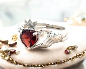 Könnte beinhalten: Ein silberner Claddagh-Ring mit einem herzförmigen, tiefroten Edelstein. Der Ring hat eine Krone über dem Herzen und Hände, die das Herz umfassen. Der Ring wird auf einer weichen, hellen Oberfläche präsentiert.