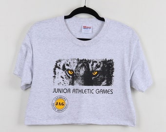 May include: Gray cropped t-shirt with a black and white graphic of a wolf's face with yellow eyes. The text "Junior Athletic Games" is printed below the graphic. A yellow circle with the text "JAG" is printed below the text.