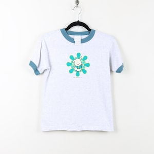 Puede incluir: Una camiseta blanca con un ribete azul. La camiseta presenta un gr&aacute;fico de un conejito blanco en un dise&ntilde;o de flor azul y verde. El texto "The Original" est&aacute; impreso debajo del gr&aacute;fico.