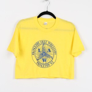 May include: Yellow cropped t-shirt with a blue and white graphic of a wheel with the text "Princeton Free Wheelers Princeton NJ".