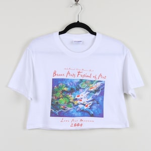 May include: White cropped t-shirt with a colorful graphic print of fish swimming in water. The text "3rd Annual Wine Festival & Beaux Arts Festival of Art Lowe Art Museum 2004" is printed on the shirt.