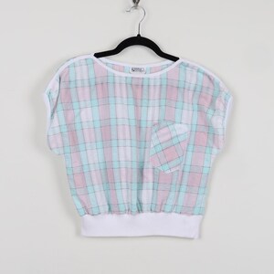 May include: A pastel plaid top with short sleeves, a white neckline, and a pocket. The top features a pattern of pink, blue, and white squares. The bottom hem is white, and the top is hanging on a black hanger.