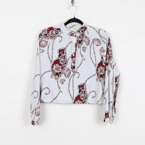 May include: A long-sleeved blouse with a white background and a repeating floral pattern in burgundy and pink. The blouse has a mandarin collar and a button-down front. The sleeves are long and end in a cuff with a burgundy floral pattern.