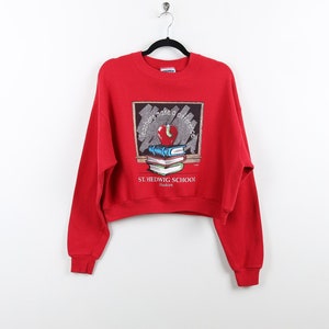 May include: Red cropped sweatshirt with a graphic of a chalkboard with the text "Teachers make a difference" and a stack of books with an apple on top. The text "St. Hedwig School" is below the graphic.