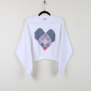 May include: White cropped sweatshirt with a large patchwork heart design in shades of pink, blue, and green.