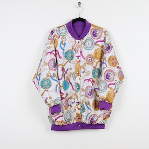 May include: A white bomber jacket with a purple lining. The jacket has a gold, pink, and blue print of chains, ribbons, and clocks.