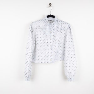 May include: A white long-sleeved button-up shirt with a small blue and red pattern. The shirt has a ruffled collar and a cropped length.