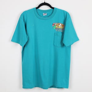Puede incluir: Camiseta turquesa con un bolsillo en el pecho izquierdo. La camiseta presenta un gr&aacute;fico colorido del logotipo de Ron Jon Surf Shop con el texto "ONE OF A KIND Cocoa Beach, FL". La camiseta est&aacute; colgada de una percha negra.