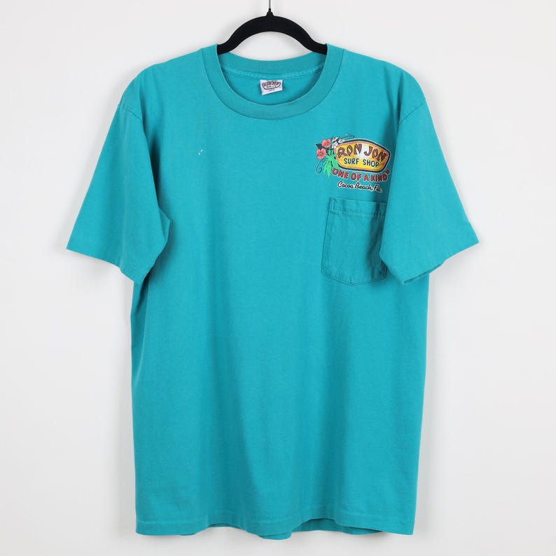 Puede incluir: Camiseta turquesa con un bolsillo en el pecho izquierdo. La camiseta presenta un gr&aacute;fico colorido del logotipo de Ron Jon Surf Shop con el texto "ONE OF A KIND Cocoa Beach, FL". La camiseta est&aacute; colgada de una percha negra.