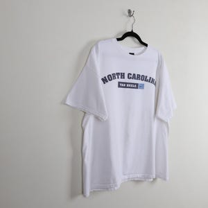 May include: White t-shirt with the words "NORTH CAROLINA" in navy blue arched across the chest. Below the text is a navy blue rectangle with "TAR HEELS" and a small blue Nike swoosh. The shirt is hanging on a black hanger.