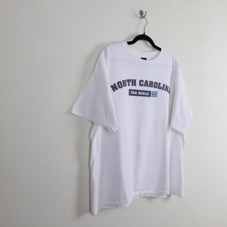 May include: White t-shirt with the words "NORTH CAROLINA" in navy blue arched across the chest. Below the text is a navy blue rectangle with "TAR HEELS" and a small blue Nike swoosh. The shirt is hanging on a black hanger.