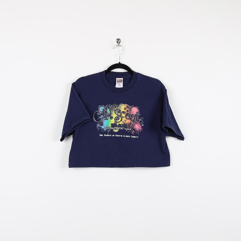 May include: A navy blue t-shirt with a colorful Girl Scouts logo and fireworks graphic. The text reads "Celebrating 90 Years Girl Scouts of Santa Clara County".