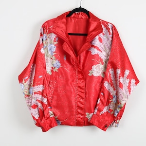May include: A red satin jacket with a floral pattern. The jacket has a full zipper, a stand-up collar, and long sleeves. The floral design includes white, blue, and purple flowers and leaves. The jacket is hanging on a black hanger.