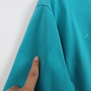 Puede incluir: Primer plano de una manga de camiseta de color turquesa. La tela parece ser una mezcla de algod&oacute;n suave. Hay unas peque&ntilde;as manchas blancas en la manga. La imagen est&aacute; bien iluminada, mostrando la textura del material.