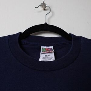May include: A navy blue t-shirt with a Fruit of the Loom tag. The tag says "100% cotton preshrunk" and "made in U.S.A. components". The tag also says "care on reverse".