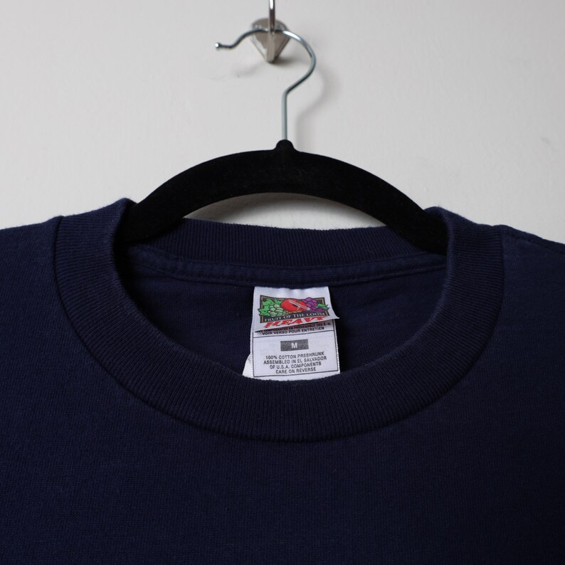 May include: A navy blue t-shirt with a Fruit of the Loom tag. The tag says "100% cotton preshrunk" and "made in U.S.A. components". The tag also says "care on reverse".