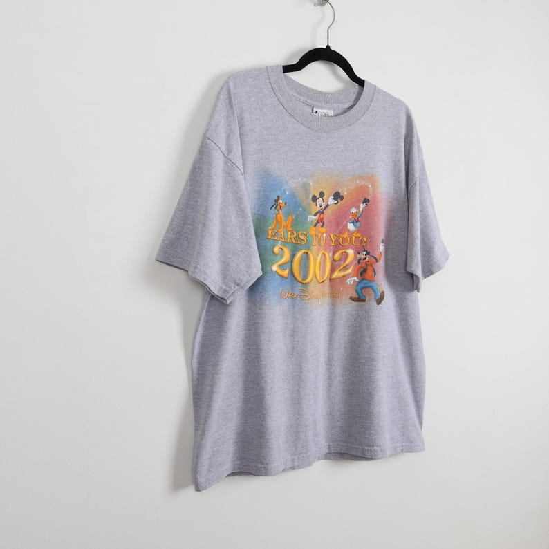Pode incluir: Uma camiseta cinza com um gr&aacute;fico colorido com personagens da Disney, incluindo Mickey Mouse e Pateta. O gr&aacute;fico inclui o texto "EARS TO YOU" e "2002". A camiseta est&aacute; pendurada em um cabide preto.