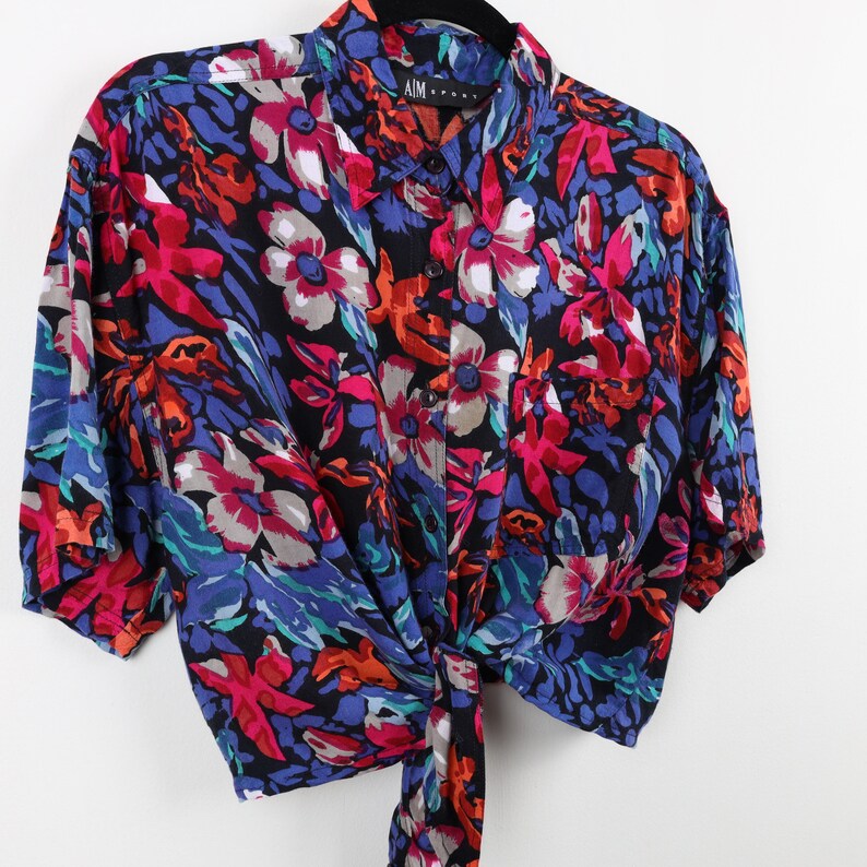 May include: A short-sleeved, cropped button-down shirt with a colorful floral print. The shirt is made of a silky fabric and has a tie at the front. The shirt is black with red, pink, blue, and orange flowers.