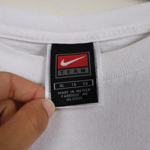 May include: Close-up of a white t-shirt collar with a black tag. The tag features a red Nike swoosh logo and the word "TEAM" in white. Below the logo are the letters "XL", "TG", and "EG". The tag reads "MADE IN MEXICO FABRIQUE AU MEXIQUE".