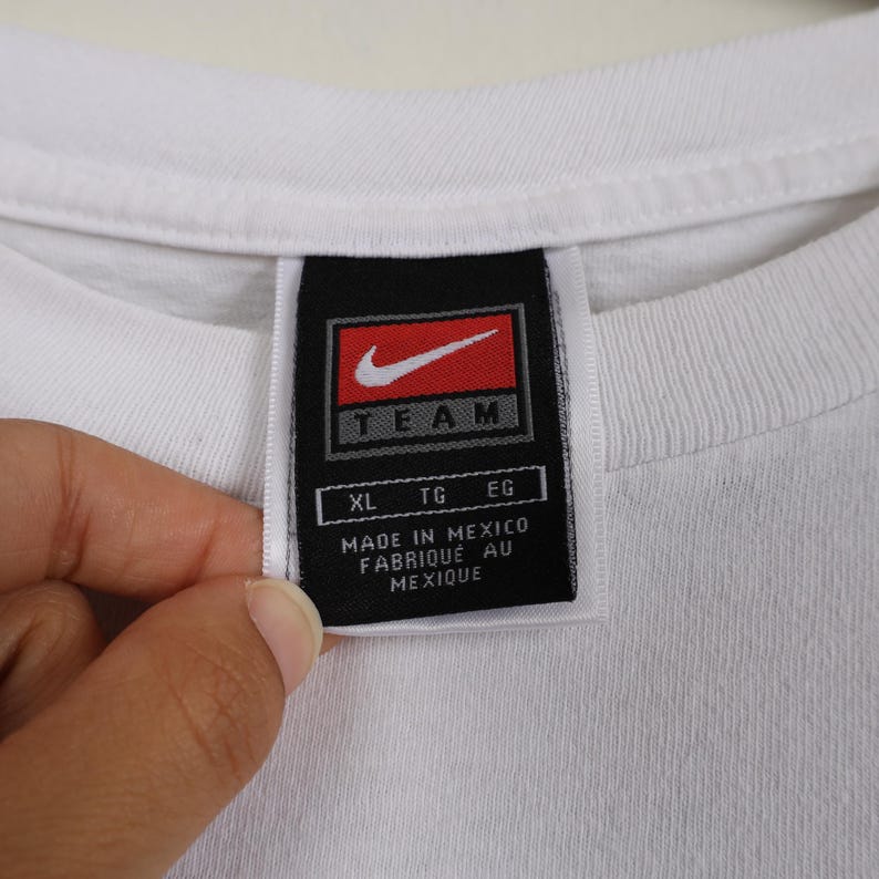 May include: Close-up of a white t-shirt collar with a black tag. The tag features a red Nike swoosh logo and the word "TEAM" in white. Below the logo are the letters "XL", "TG", and "EG". The tag reads "MADE IN MEXICO FABRIQUE AU MEXIQUE".