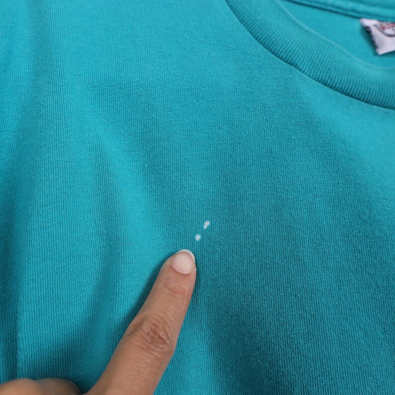 Puede incluir: Primer plano de una camiseta de color turquesa. La tela tiene un aspecto texturizado. Un dedo se&ntilde;ala dos peque&ntilde;as manchas blancas en la camiseta. El cuello de la camiseta es visible en la esquina superior derecha.