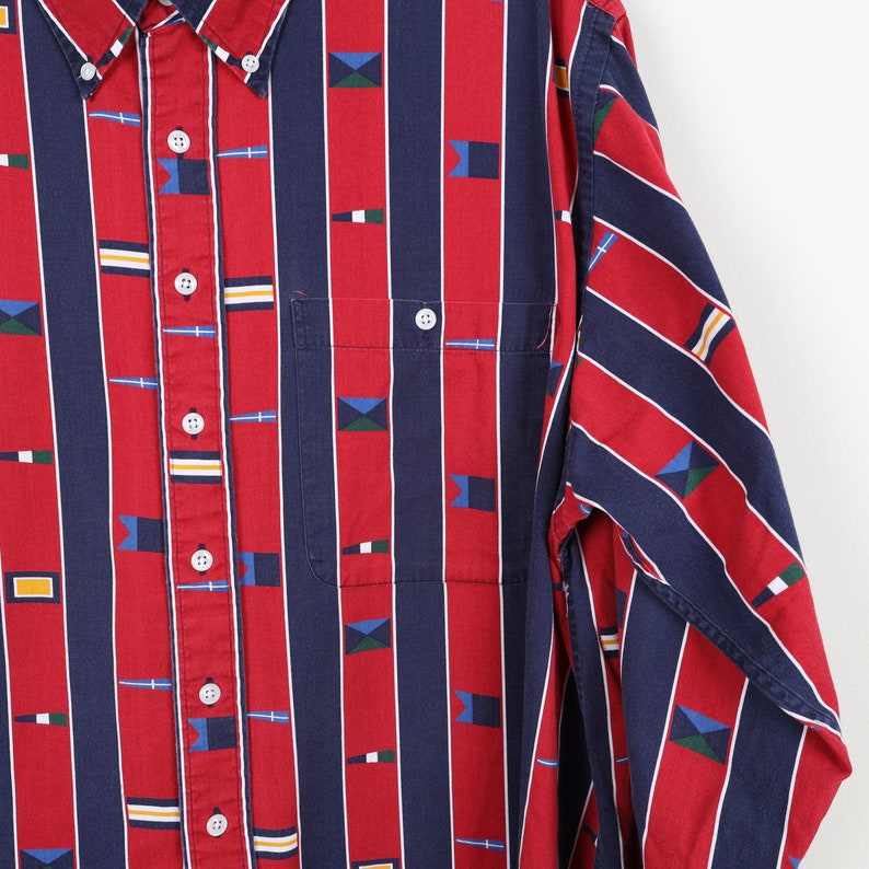 Puede incluir: Una camisa de manga larga con botones, a rayas rojas y azules, con un patr&oacute;n de peque&ntilde;as banderas y s&iacute;mbolos n&aacute;uticos.
