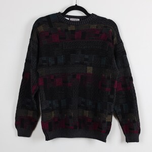 May include: A dark gray sweater with a geometric pattern in shades of burgundy, teal, and gold. The sweater has a crew neck and long sleeves, and is hanging on a black hanger against a white background.